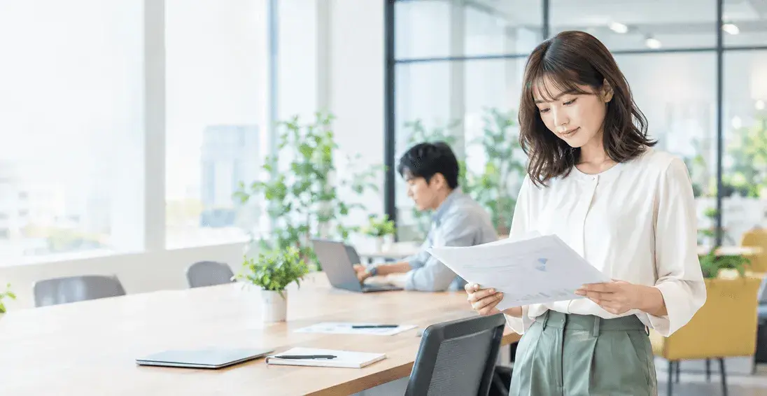 総務担当の女性が、オフィスのデスクでノートPCの管理表を確認している。自然光。