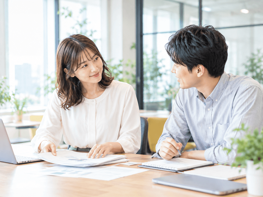同じ女性が、同じオフィスのデスクで、笑顔でノートPCを閉じて一息ついている。自然光。
